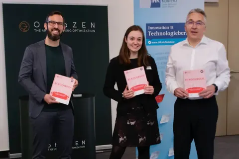 Simon Tiffert with two guests of the AI Innovation Camp in front of the OptaZEN banner and the IHK Koblenz stand